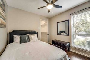 Quarto com cama, ventoinha no teto, um banco, guarda-roupa e janela com persianas.