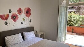 A bedroom with a large bed, a nightstand, a wall-mounted lamp, and a window with a view of greenery outside. - Casa giardino San Pietro (Roma)