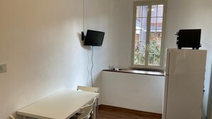 A small, clean room with a white table, two chairs, a white refrigerator, a window with a view of buildings, and a flat-screen TV mounted on the wall. - Casa giardino San Pietro (Roma)