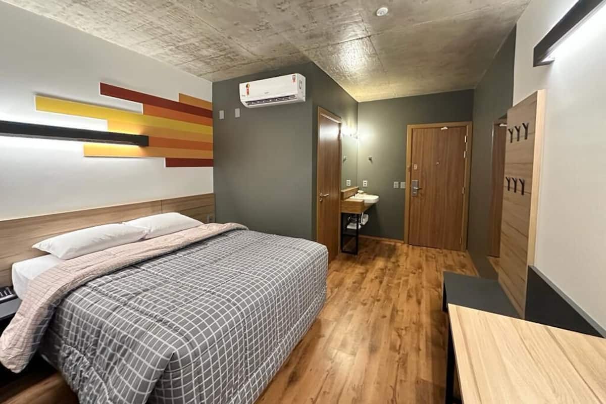 Una habitación de hotel con piso de madera, una cama con colcha a cuadros, aire acondicionado de pared, un armario de madera y un baño pequeño con lavamanos y espejo.