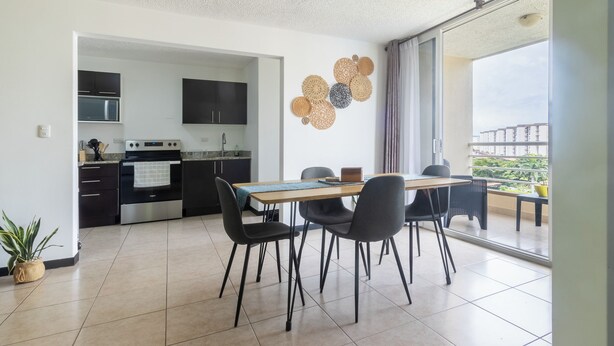 Une cuisine moderne avec des meubles de couleur sombre, un four à micro-ondes et une cuisinière. On y trouve un coin repas avec une table et des chaises, ainsi qu’une grande fenêtre offrant une vue sur les bâtiments et la verdure.