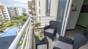 Un balcon avec deux fauteuils en osier, une petite table et une plante en pot. On aperçoit une piscine à l’arrière-plan.