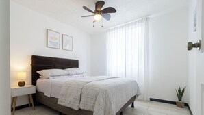 Une chambre à coucher avec un lit, une table de chevet avec une lampe, un ventilateur de plafond et une fenêtre avec des rideaux.
