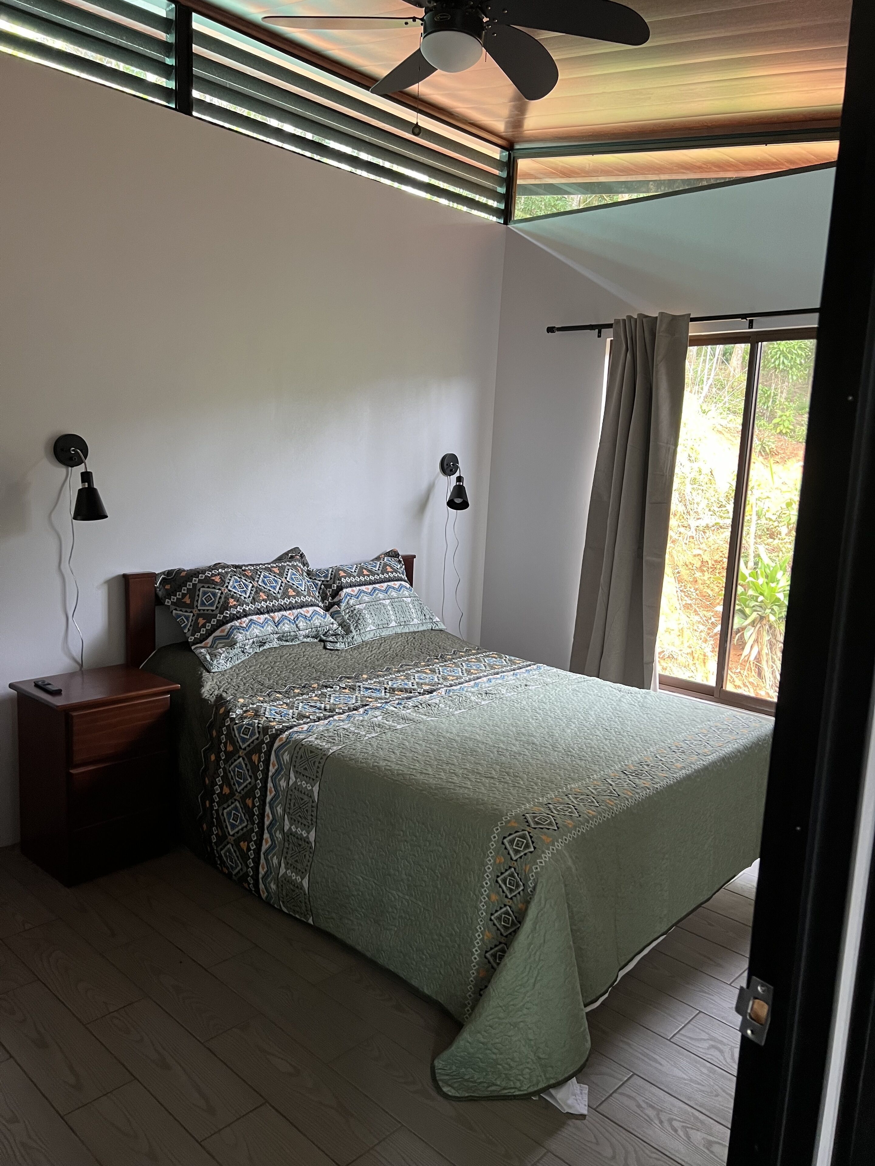 A bedroom with a bed, bedside lamps, a ceiling fan, a window with curtains, and a nightstand.