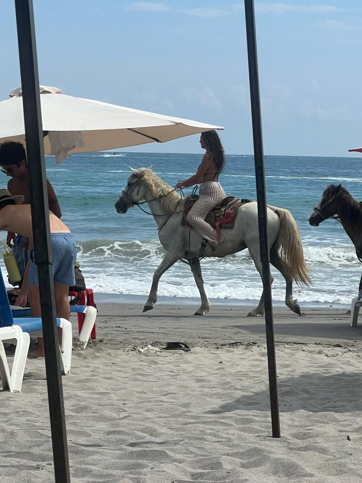 En badestrand med en person, der rider på en hvid hest, en anden hest i baggrunden, samt strandstole og parasoller.