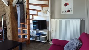 A living room with a wooden spiral staircase, a flat-screen TV, a red sofa, and a radiator. - Joli Gîte Rural Pour un Séjour à la Campagne (DIEULIVOL)