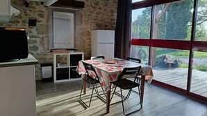 A dining area with a table set for two, a white refrigerator, and a stone wall. - Joli Gîte Rural Pour un Séjour à la Campagne (DIEULIVOL)