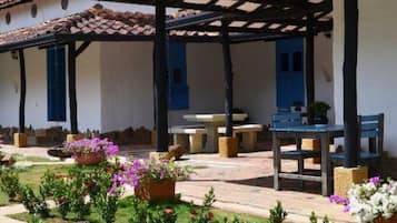 A covered outdoor area with tables and chairs, surrounded by a garden with small plants and flowers.