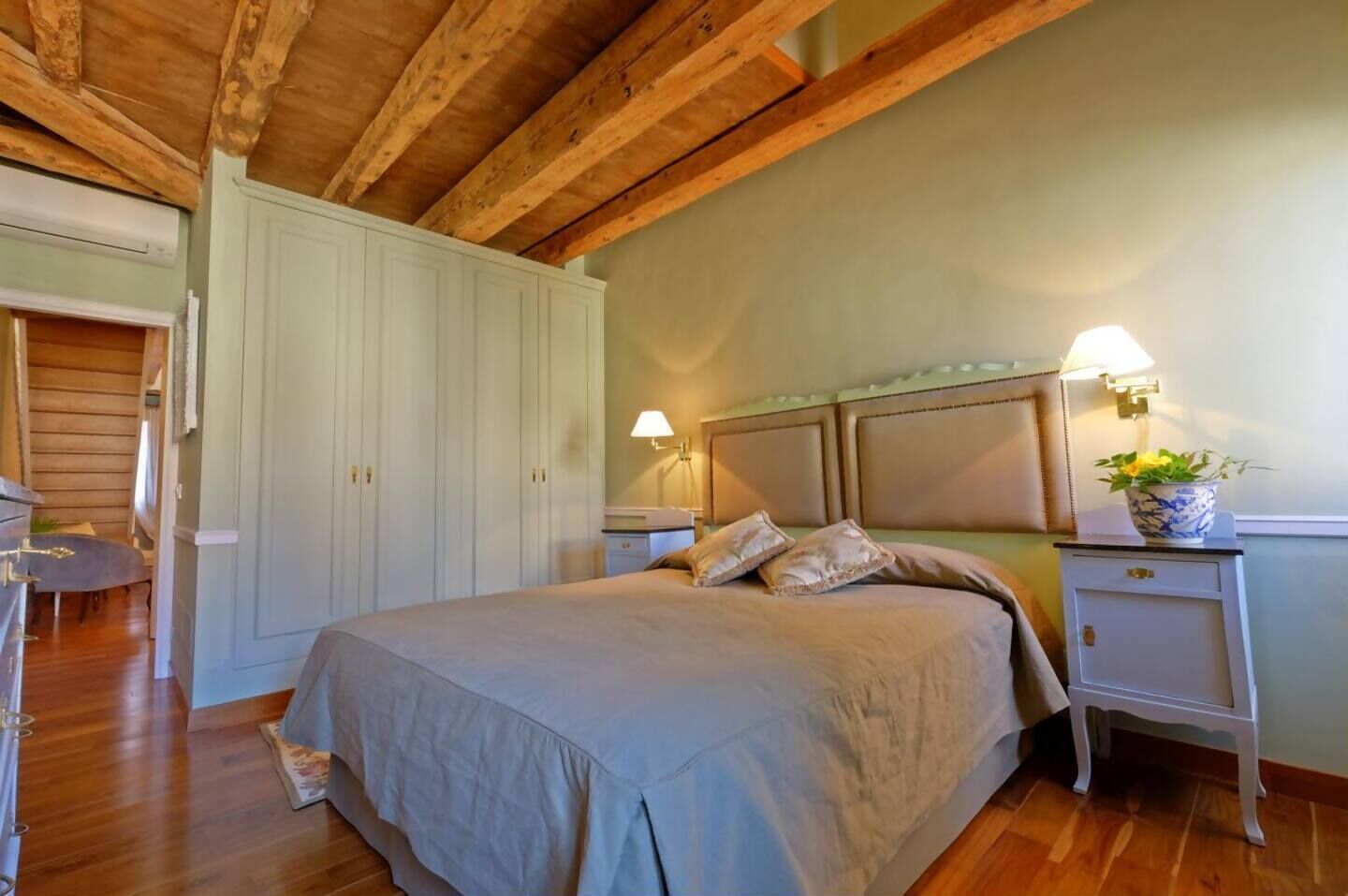 A bedroom with a bed, wooden ceiling beams, a nightstand with a lamp, and a wardrobe.