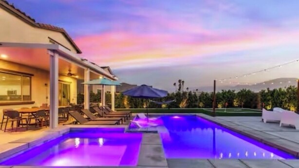 A swimming pool with a modern design, surrounded by a covered patio area with seating and a view of the landscape. - The Chateau- Pool, Spa, BBQ, Fire pit, Sleeps 19 (Coachella)
