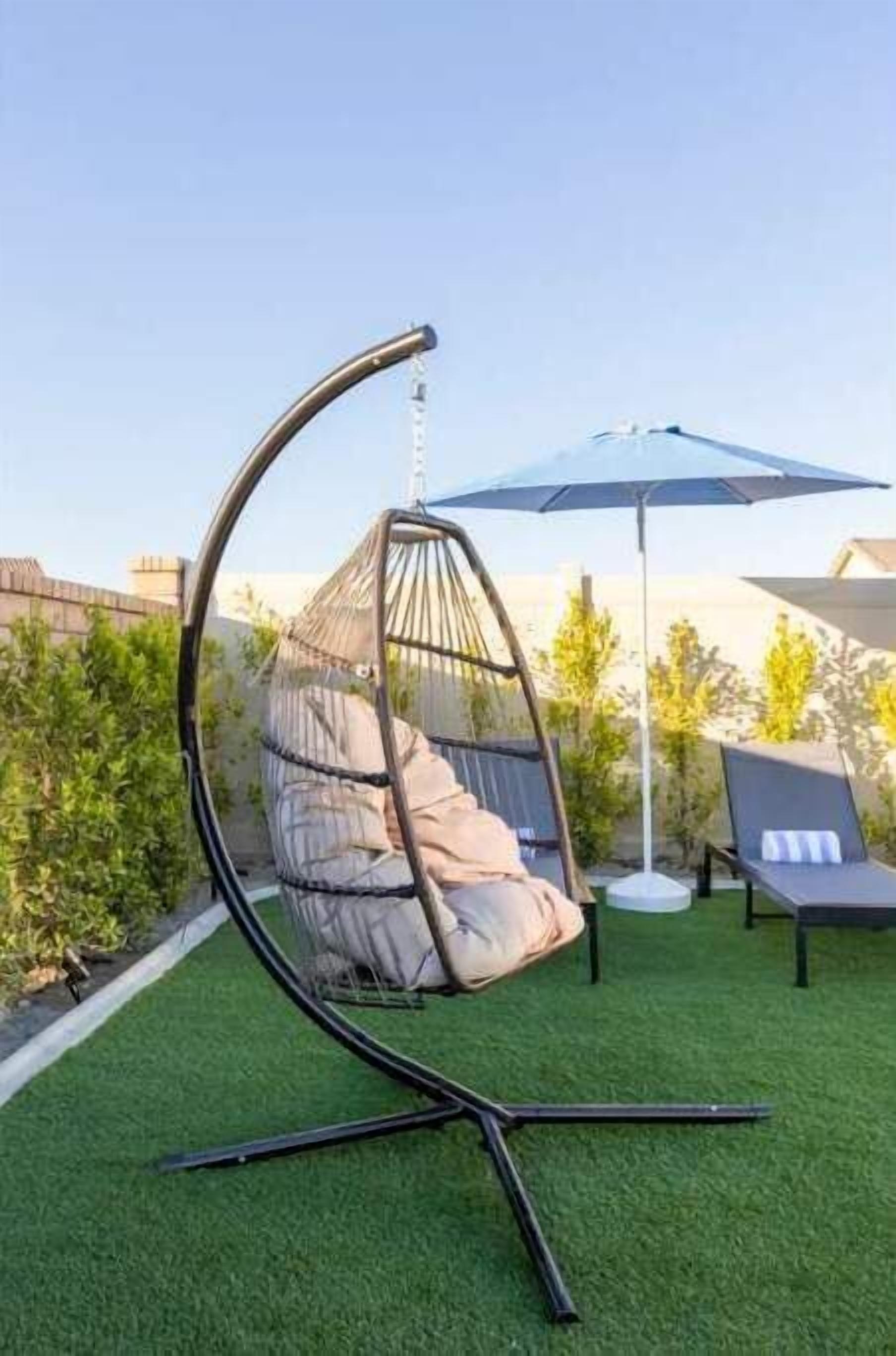 En hængekøje med puder på et stativ, et rundt bord og en blå parasol i en have.