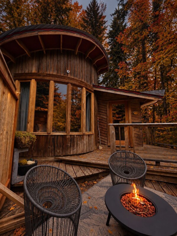 A wooden cabin with a circular deck, two wicker chairs, and a central fire pit.