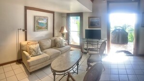 A living room with a sofa, a coffee table, and a TV. There are two chairs and a lamp. The room has a large window and a door leading outside.