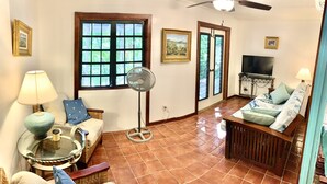 A living room with a sofa, a small table with a lamp, a fan, and a TV. There are two windows and a door. - Azul La Mer - Aqua Bay Villas (Cruz Bay)