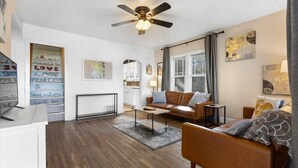 Una sala de estar moderna con un sofá de cuero marrón, una mesa de centro y un ventilador de techo. Hay una ventana con cortinas, una chimenea y una pared con citas motivacionales.
