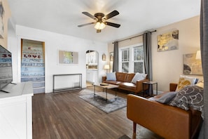A modern living room with a brown leather sofa, a coffee table, and a ceiling fan. There is a window with curtains, a fireplace, and a wall with motivational quotes.