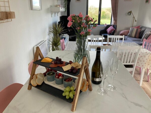 A dining area with a marble table set for a meal, featuring a cheese board, grapes, olives, and bread. A bottle of wine and glasses are also visible.