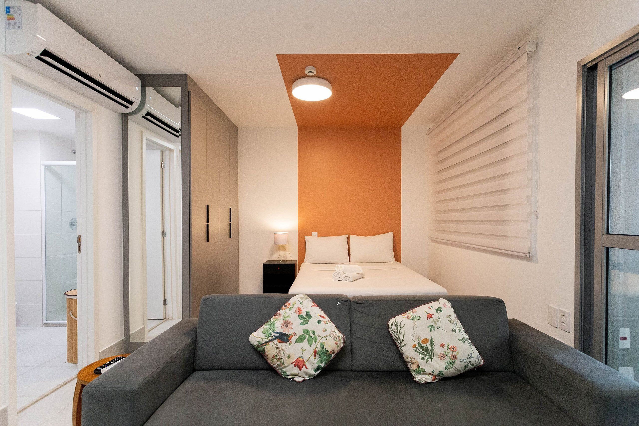 Una habitación de hotel moderna con un sofá gris, dos almohadas estampadas, una cama con ropa de cama blanca, un equipo de aire acondicionado, una lámpara de noche y una ventana con persianas.