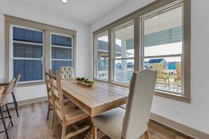 Un espace repas comprenant une table et des chaises en bois, de grandes fenêtres équipées de stores et une vue sur d’autres bâtiments.