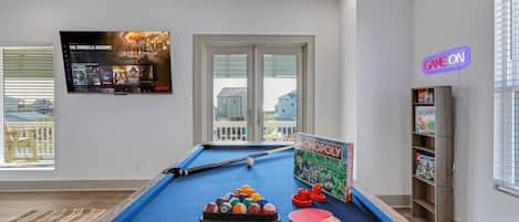 Een biljart met een keu, ballen en een Monopoly spel in een kamer met een groot raam, een neonbord en een boekenkast.