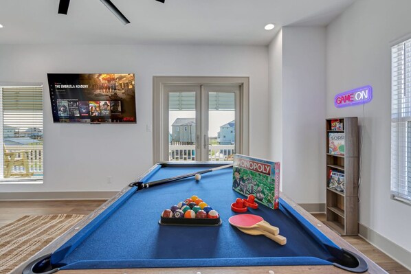 Une table de billard avec une queue de billard, des billes et un jeu de société Monopoly dans une pièce dotée d'une grande fenêtre, d'un néon et d'une bibliothèque.
