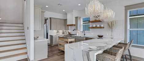 A modern kitchen with a marble island, wooden flooring, and a unique chandelier.