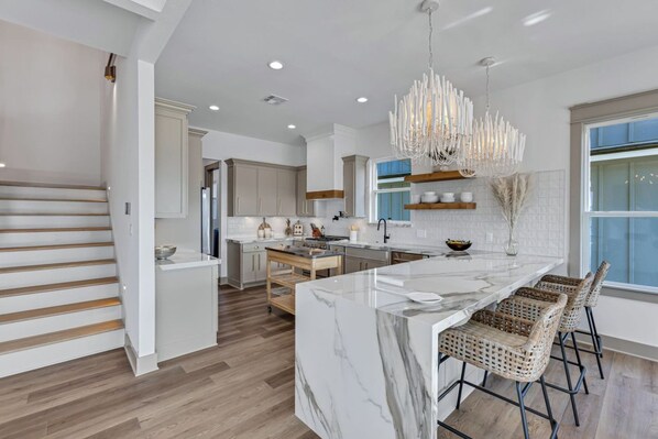A modern kitchen with a marble island, wooden flooring, and a unique chandelier.