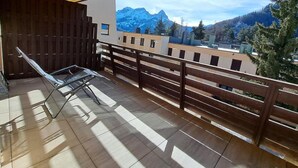En balkon med metalseng, trægelænder og udsigt til bjerge og bygninger.