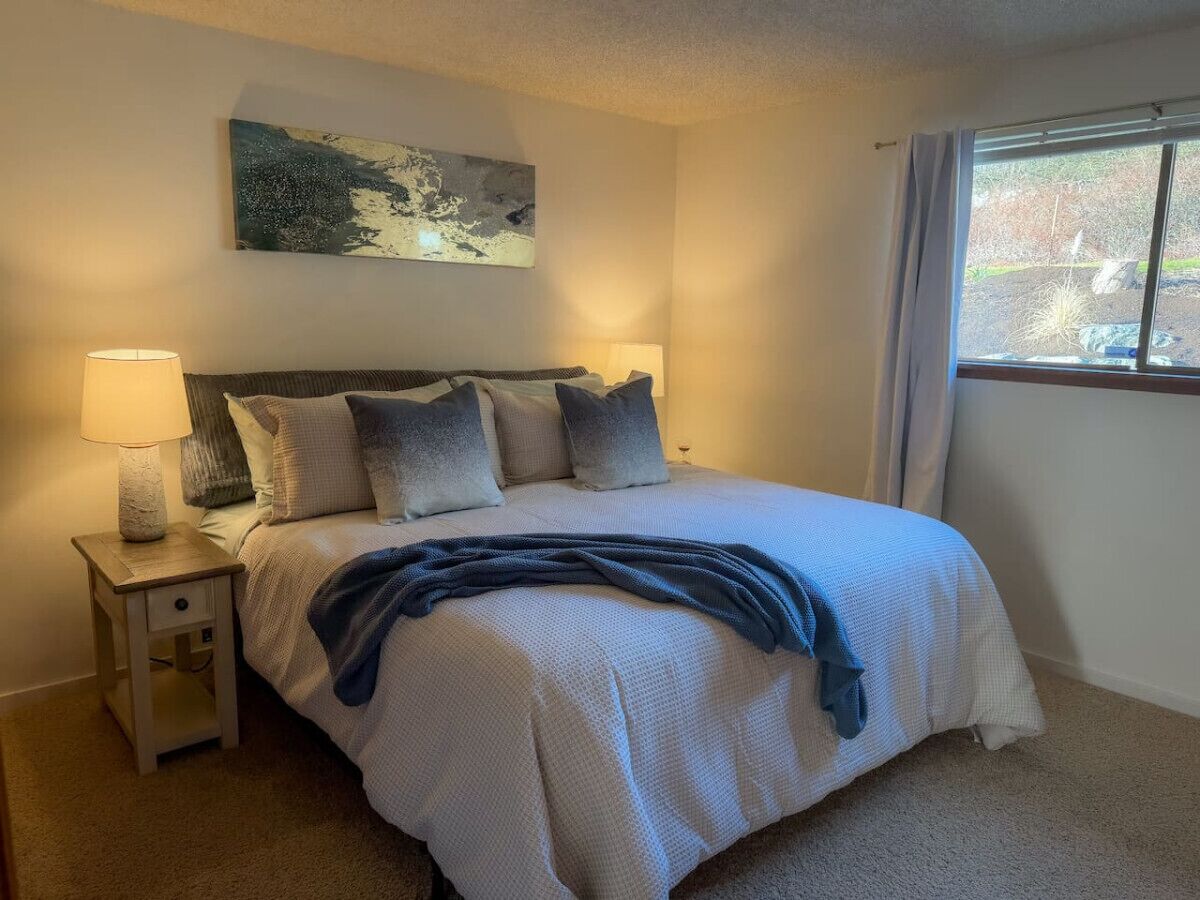 A bedroom with a bed, a nightstand with a lamp, a painting on the wall, and a window with curtains.