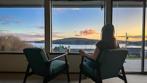 Une pièce avec deux fauteuils placés face à une grande fenêtre donnant sur une étendue d’eau et un paysage lointain, au moment du coucher du soleil.
