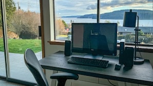 Un bureau d’ordinateur équipé d’un écran, d’un clavier et de haut-parleurs, placé près d’une fenêtre donnant sur un lac et des collines.
