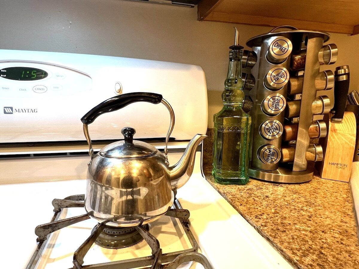 Una tetera de acero inoxidable sobre una cocina de gas portátil, una botella verde y un juego de cuchillos de cocina sobre una encimera de granito.