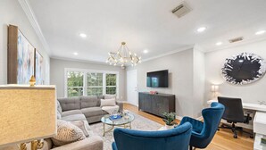 A modern living room with a grey sofa, blue armchairs, a glass coffee table, and a TV on the wall. - The Queen’s Gambit II (Houston)