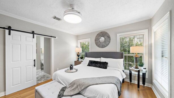 A bedroom with a large bed, a bedside table, a lamp, a decorative wall piece, and a window with blinds. - The Queen’s Gambit II (Houston)