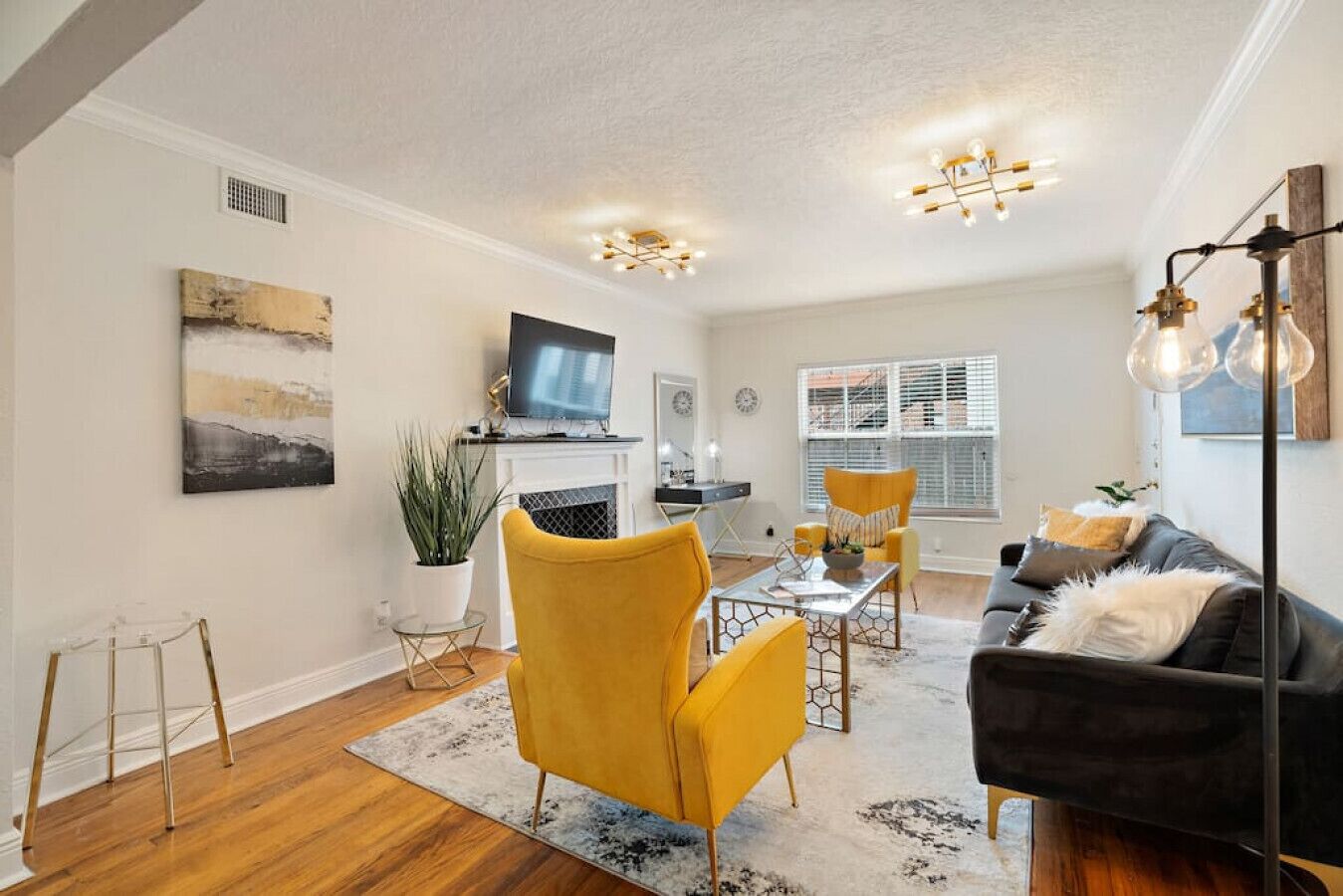 A living room with a fireplace, yellow chairs, a black sofa, and a large window with blinds.