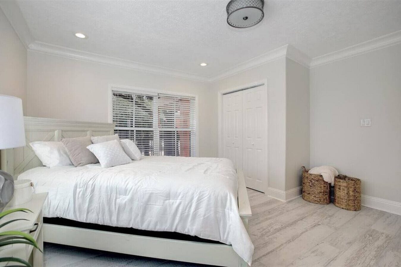A bedroom with a bed, white walls, a window with blinds, a door, and two wicker baskets.
