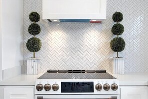 A kitchen with a white herringbone backsplash, a central range hood, and two potted topiary plants. - The Bishop's Haven 2 (Houston)