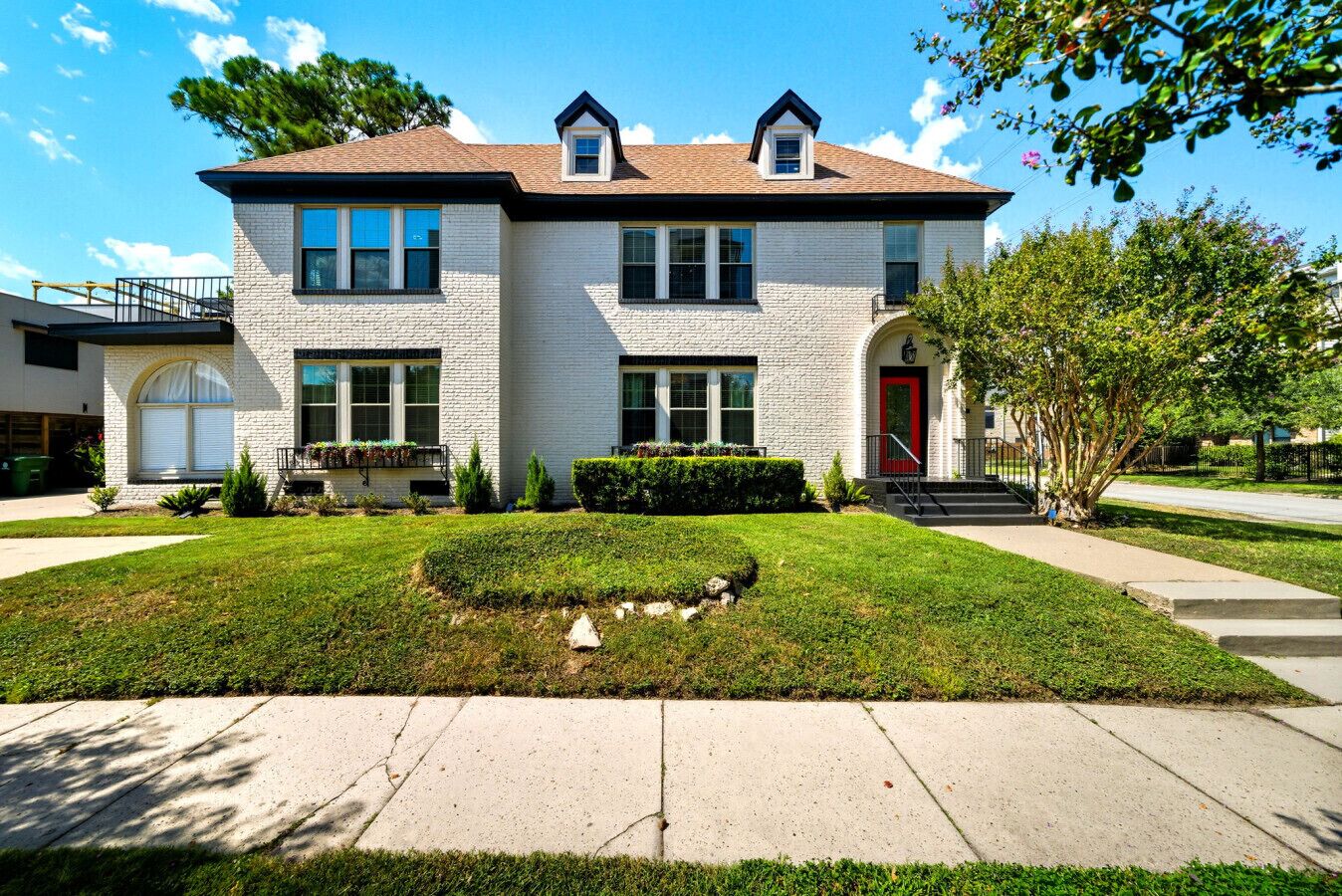 A two-story house with a red door, a small front yard with a hedge, and a clear blue sky.