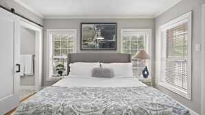 A bedroom with a large bed, two windows with blinds, a painting above the bed, and a nightstand with a lamp. - The Queen's Gambit I (Houston)