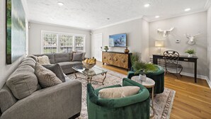 A living room with a gray sofa, a green armchair, a coffee table, and a wooden cabinet. There is a painting on the wall and a plant on a side table. - The Queen's Gambit I (Houston)