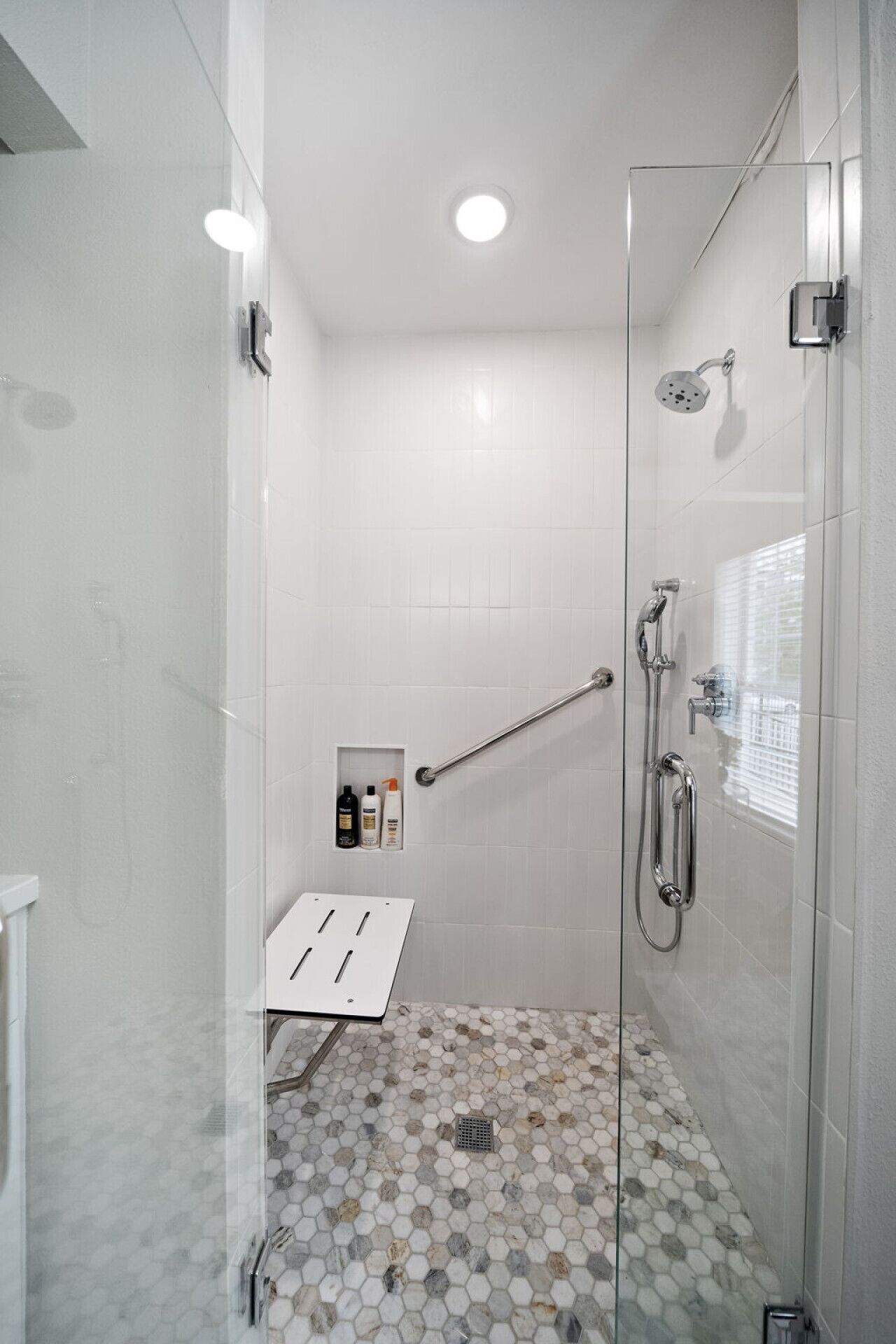 A modern shower room with a glass door, white tiled walls, a hexagonal tile floor, and a handrail.