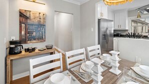 A modern dining area with a wooden table set for four, white chairs, and a kitchen in the background. - The Queen's Gambit I (Houston)