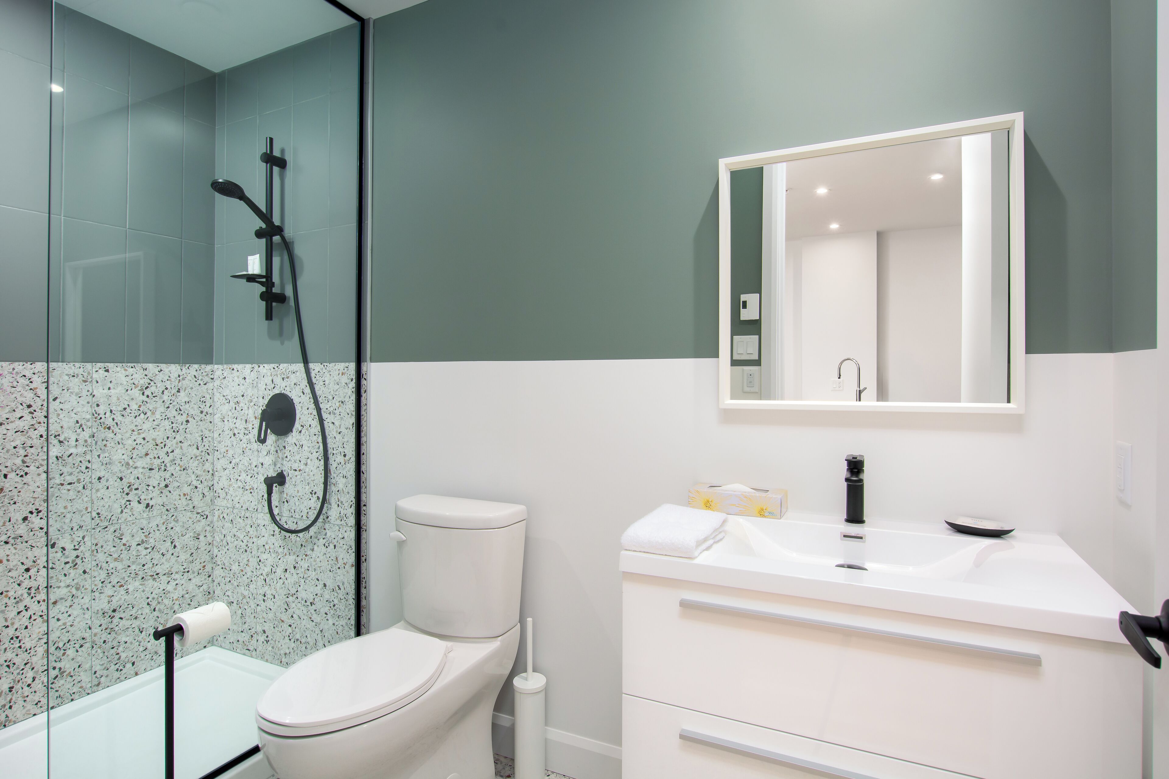 A modern bathroom with a glass shower enclosure, a white toilet, a white sink with a mirror above it, and a white vanity with drawers.