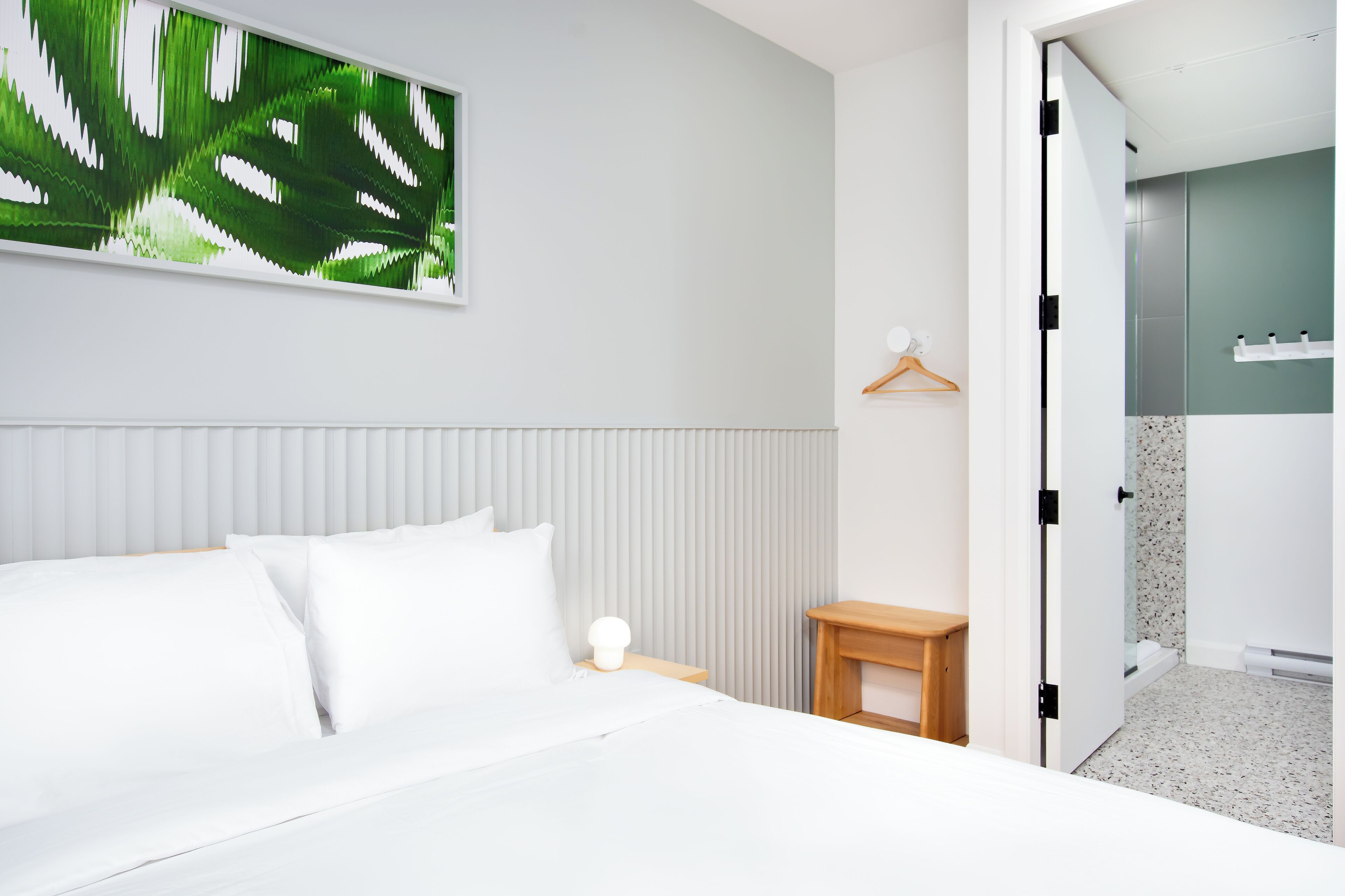 A modern hotel room with a neatly made bed, a bedside table with a lamp, a wall-mounted artwork, and a view into the bathroom.