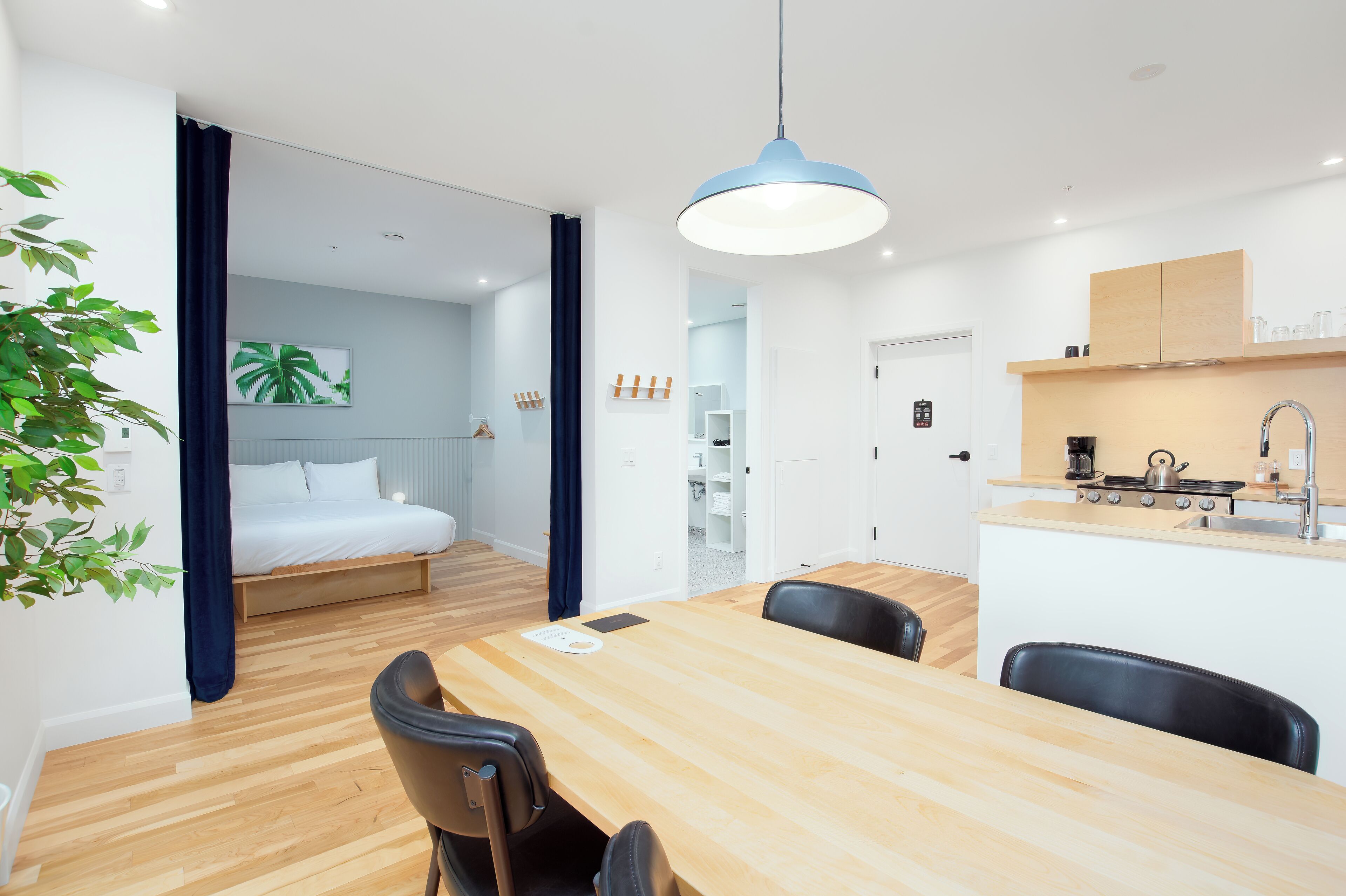 A modern hotel room with a wooden dining table, black chairs, a kitchenette, a bed with white linens, and a large window.