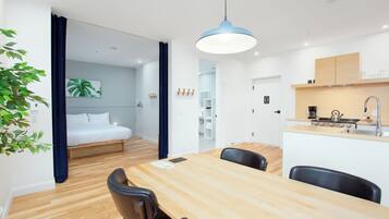 A modern hotel room with a wooden dining table, black chairs, a kitchenette, a bed with white linens, and a large window.