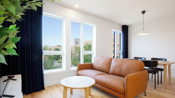 Un salon moderne avec un canapé en cuir, une table basse ronde en bois et un coin repas comprenant une petite table et des chaises. De grandes fenêtres offrent une vue sur le paysage urbain.
