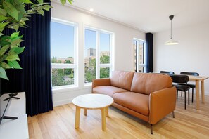 A modern living room with a leather sofa, a round wooden coffee table, and a dining area with a small table and chairs. Large windows provide a view of the cityscape. - Les Lofts de Vitré - Par Les Lofts Vieux-Québec (Québec City)