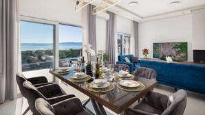 Un espace repas moderne avec une table en verre pouvant accueillir six personnes, un canapé bleu et une grande fenêtre donnant sur la mer.
