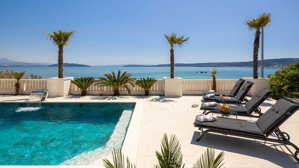 Un espace aménagé au bord de la piscine, avec des chaises longues et des palmiers, offrant une vue sur une mer d’un bleu limpide et sur des montagnes lointaines.